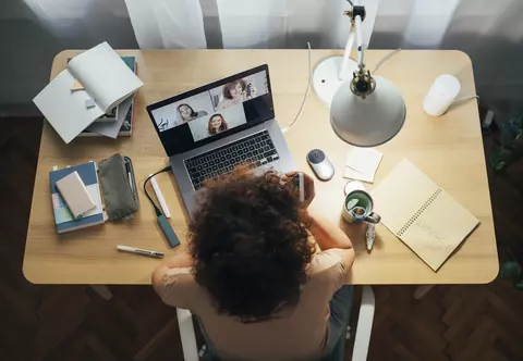 Mulher sentada próximo a uma mesa com vários objetos sobre ela. A mulher está utilizando o notebook, fazendo uma reunião virtual com outras três pessoas.