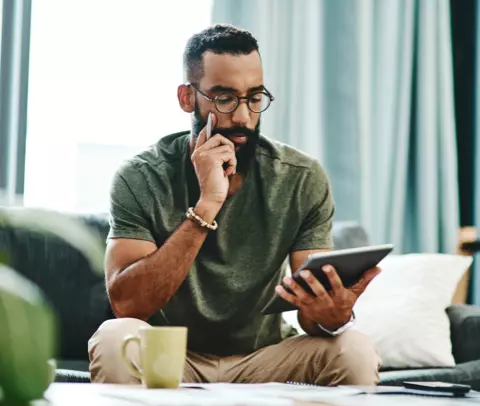 Homem de camiseta verde está sentado em um sofá e segura o rosto com a mão. Na mesma mão está uma caneta. Ele olha concentrado para um tablet que está em sua outra mão. Na sua frente tem uma mesa de centro, com papéis, celular e uma caneca.