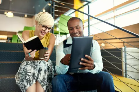  Imagem de uma mulher e um homem sorrindo e sentados em uma escada de metal. Ela segura um caderno e um copo de café, enquanto olha para o tablet que ele está segurando.